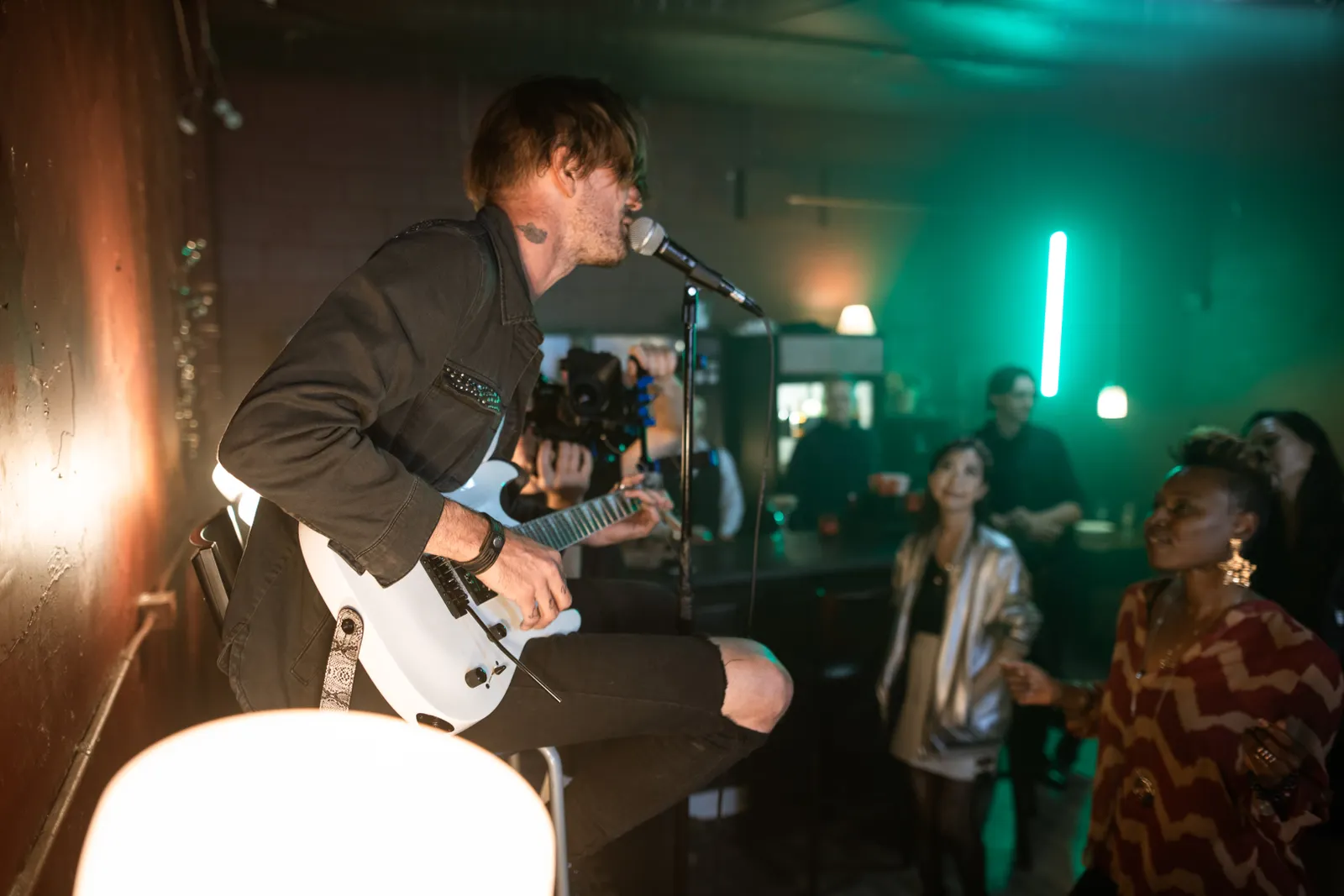 A singer-guitarist mid-song under warm and green stage light, the crowd close enough to touch